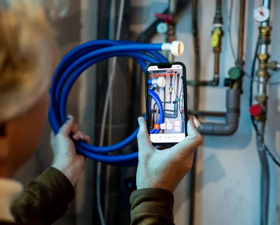 Iemand houdt een smartphone voor een technische installatie met leidingen en meters. Op het scherm wordt zichtbaar een foto gemaakt van de situatie. Het beeld toont een moment waarop iemand documentatie vastlegt van een technische omgeving.
