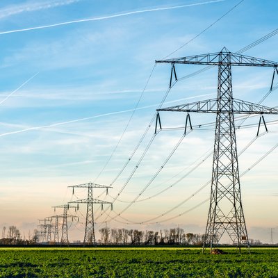 Een rij hoogspanningsmasten met elektrische kabels strekt zich uit over een groen veld. De lucht is blauw met enkele wolken en vliegtuigstrepen. Op de achtergrond staan bomen en een lichte gloed van de ondergaande zon.