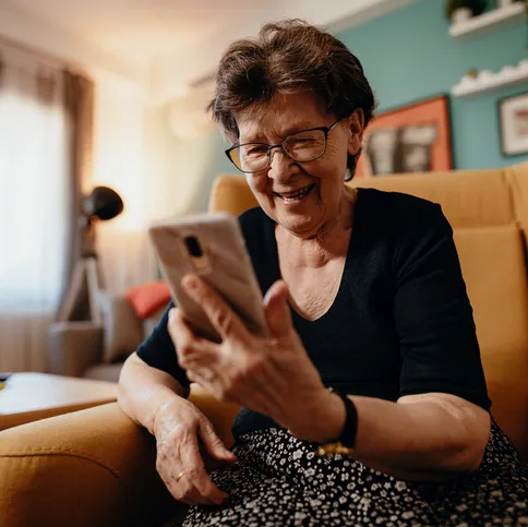 Een oudere vrouw met bril en zwarte kleding zit in een gele fauteuil en lacht terwijl ze haar smartphone vasthoudt. De achtergrond toont een sfeervolle woonkamer met decoraties, een bank en een raam met gordijnen