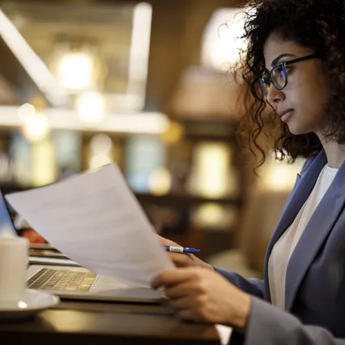 Een vrouw met krullend haar, bril en een blauw pak leest aandachtig een document terwijl ze een pen vasthoudt. Ze zit aan een tafel met een laptop en een koffiekopje in een sfeervol café met warme verlichting