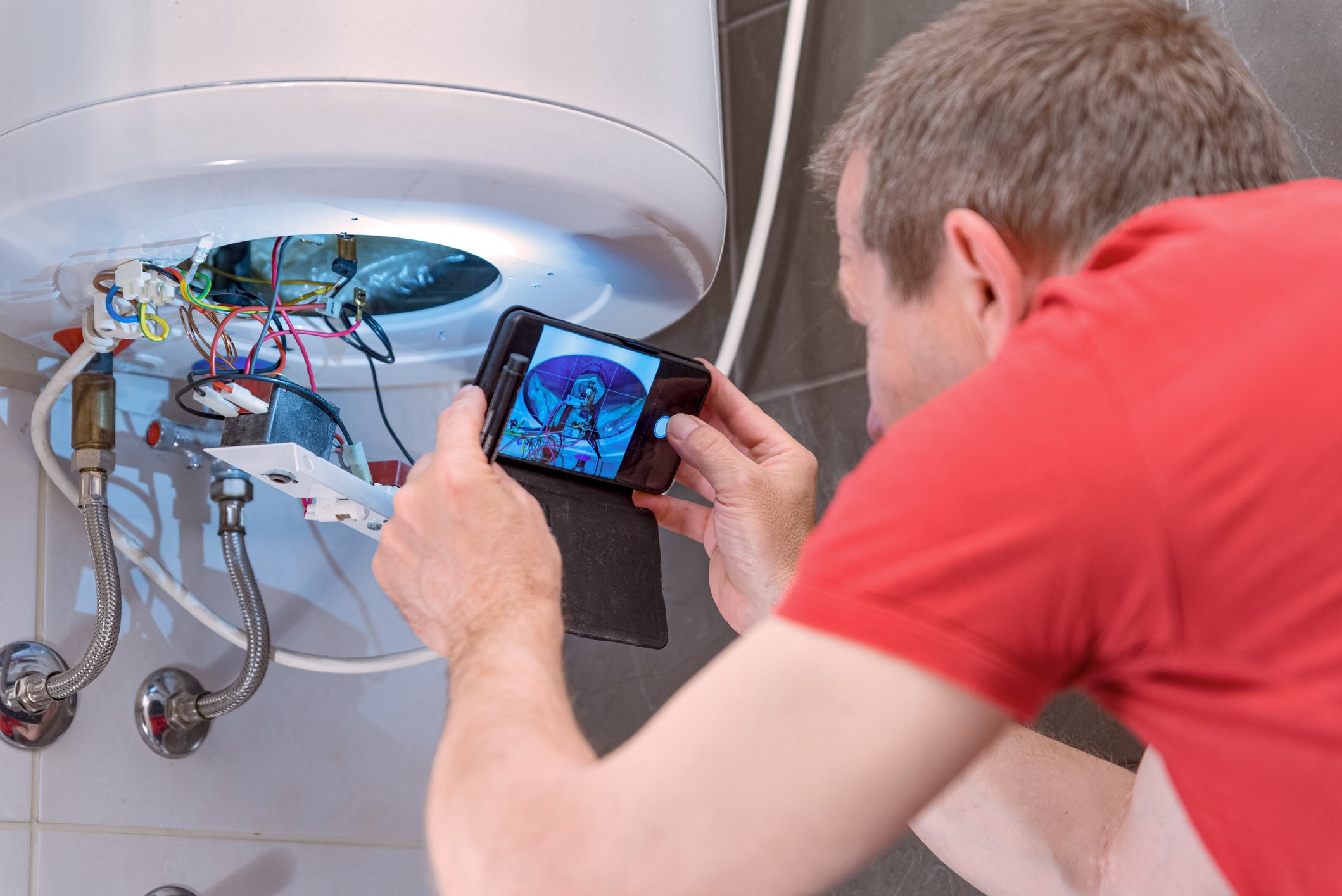 Een man in een rood shirt fotografeert de bedrading van een geopende boiler met zijn smartphone. De boiler hangt aan een betegelde muur, met blootliggende draden en aansluitingen zichtbaar. De telefoon toont een referentieafbeelding voor de reparatie.