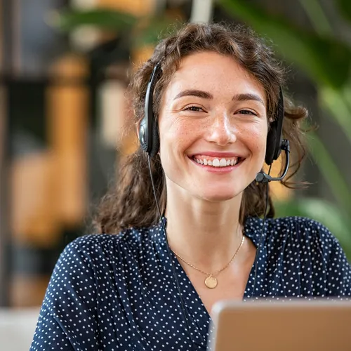 Een vrouw met krullend haar, een headset en een donkerblauwe blouse met stippen lacht terwijl ze achter haar laptop zit. De achtergrond is modern ingericht met planten en warm licht