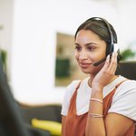 Een jonge vrouw met een headset zit geconcentreerd achter een computerscherm in een moderne, lichte kantooromgeving. Ze glimlacht vriendelijk terwijl ze luistert of spreekt. Haar outfit is casual en stijlvol; de sfeer professioneel en klantgericht.