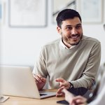 Een man in een lichtgekleurde trui zit achter een laptop en voert een vriendelijk gesprek met een collega. Hij lacht en gebaart betrokken, in een lichte kantoorruimte met moderne inrichting. De sfeer is open en professioneel.
