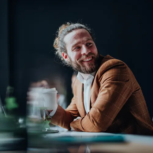 Een man met een lichtbruine blazer en witte blouse zit glimlachend aan een tafel in een modern kantoor. Hij houdt een koffiebeker vast en kijkt ontspannen opzij. De sfeer is informeel en warm, met zachte focus op de voorgrond en donkere achtergrond.