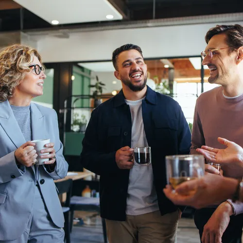 Vier collega’s staan samen in een kantoor en lachen tijdens een informeel gesprek. Ze houden kopjes koffie of thee vast. De vrouw links draagt een lichtgrijs pak en bril, de anderen dragen een donkerblauwe jas, een beige trui en een wit gestreepte blouse.