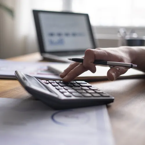 Een persoon in een blauw overhemd typt op een rekenmachine terwijl hij een pen vasthoudt. Op het houten bureau liggen financiële documenten, een laptop en een koffiebeker. Het scherm van de laptop toont een rapport met grafieken.