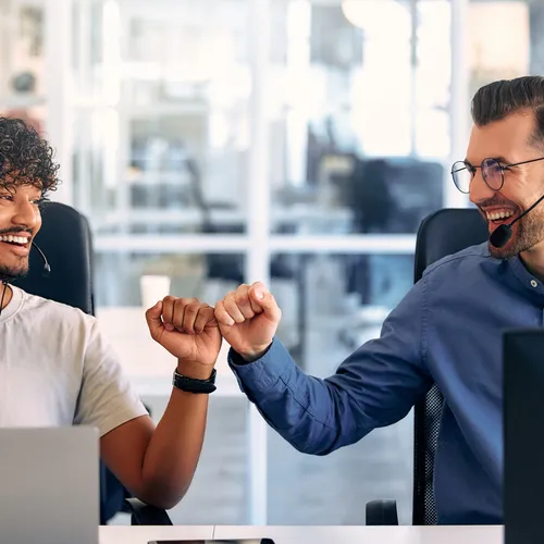 Twee jonge mannen met headsets zitten achter computers in een moderne klantenservice en geven elkaar een fist bump. Ze lachen en kijken naar elkaar. De één draagt een licht T-shirt, de ander een blauw overhemd. De ruimte heeft glazen wanden en een open ka