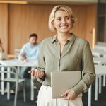 Een vrouw met blond haar en een olijfgroene blouse staat glimlachend in een vergaderruimte. Ze houdt een gesloten laptop vast en lijkt klaar voor een presentatie of gesprek. Op de achtergrond overleggen collega’s informeel aan een tafel.