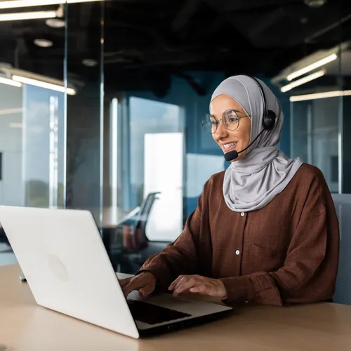 Een vrouw met een hoofddoek, bril en een headset werkt op een laptop in een kantoor met glazen wanden. Ze lacht tijdens een gesprek en typt. De moderne werkruimte heeft een rustige sfeer met neutrale kleuren en moderne plafondverlichting.