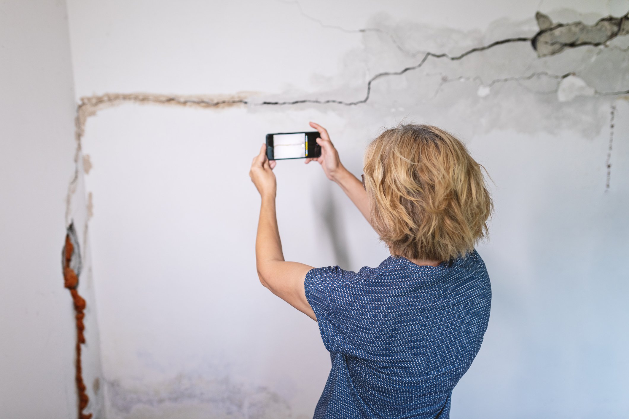 Een vrouw met blond haar en een blauwe blouse maakt een foto van een muur met grote scheuren en loslatend pleisterwerk. Ze houdt haar smartphone omhoog en staat met haar rug naar de camera in een beschadigde kamer.