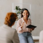 Een vrouw met een gestreepte blouse en tablet in de hand spreekt enthousiast tijdens een overleg. Ze zit op tafel en richt zich tot twee collega’s in een lichte, moderne ruimte. De sfeer is informeel, betrokken en professioneel.