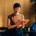 Een vrouw met een korte pony en een bruine top staat glimlachend met een feloranje tablet in haar handen. Ze leest of presenteert iets in een moderne werkomgeving. Op de achtergrond zit een collega achter een laptop, in een rustige, warme setting.