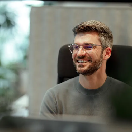 Een jonge man met een grijze trui en bril zit glimlachend op een bureaustoel in een moderne kantooromgeving. Hij kijkt opzij, zichtbaar ontspannen en tevreden. De achtergrond is licht en onscherp, met planten en akoestische panelen in beeld.
