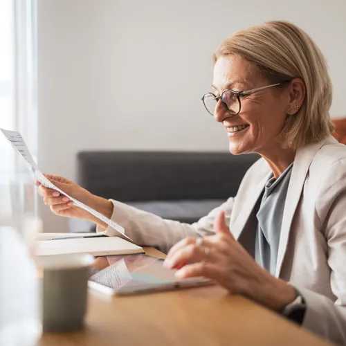 Een vrouw met blond haar, bril en een beige blazer zit aan een houten tafel en bekijkt lachend een document. Op tafel liggen een tablet, notitieboek en een koffiekopje. De achtergrond toont een bank met kussens in een lichte woonkamer