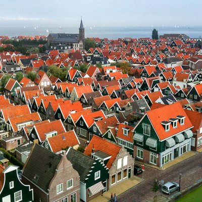 Een Nederlands dorp met traditionele huizen met rode dakpannen en groene gevels. De smalle straten en grachten lopen door de wijk. Op de achtergrond staat een kerk met een toren en is water zichtbaar met zeilboten in de verte.