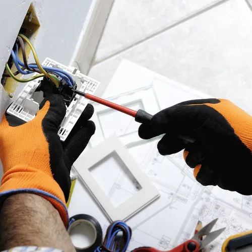 Een elektricien met oranje werkhandschoenen gebruikt een schroevendraaier om een stopcontact te installeren. De muur heeft open bedrading met blauwe, gele en groene draden. Op de vloer liggen elektrische gereedschappen, isolatietape en bouwtekeningen